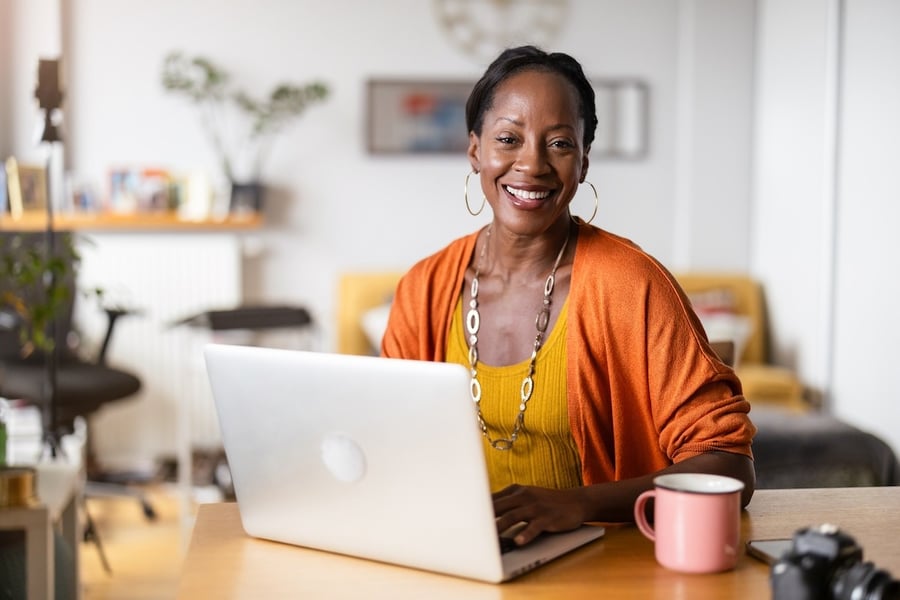 A smiling woman sits at a desk with a laptop and a pink coffee mug in a cozy home setting.